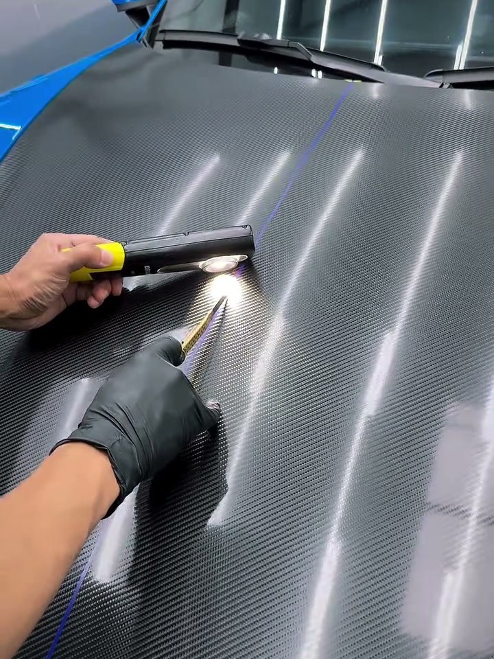 Gloved hand guiding a squeegee across carbon fiber under a work lamp